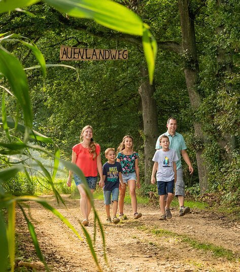 Eine Familie mit drei Kindern läuft unter dem Schild durch, das an einem Baum hängt und auf dem Auenlandweg steht: Der Eingang zu einem der beliebtesten Erlebniswege Sieg.