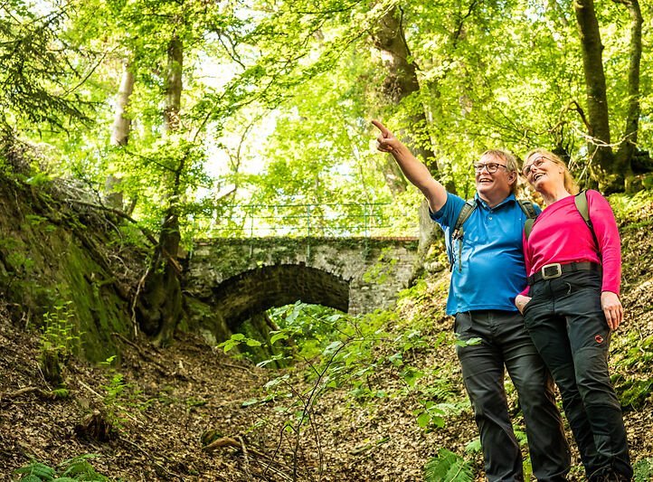 Ein Wanderer-Paar steht glücklich mit Rucksack im Wald und lacht. Der Mann zeigt auf etwas. Im Hintergrund erkennt man eine Brücke zwischen den Bäumen. 