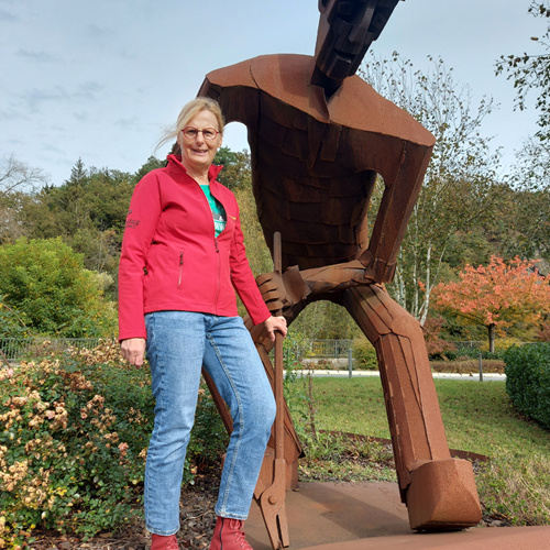 Stadtführerin Christiane Weber steht vor einem Denkmal und trägt eine rote Stadtführerjacke mit dem Schriftzug wisserland.de